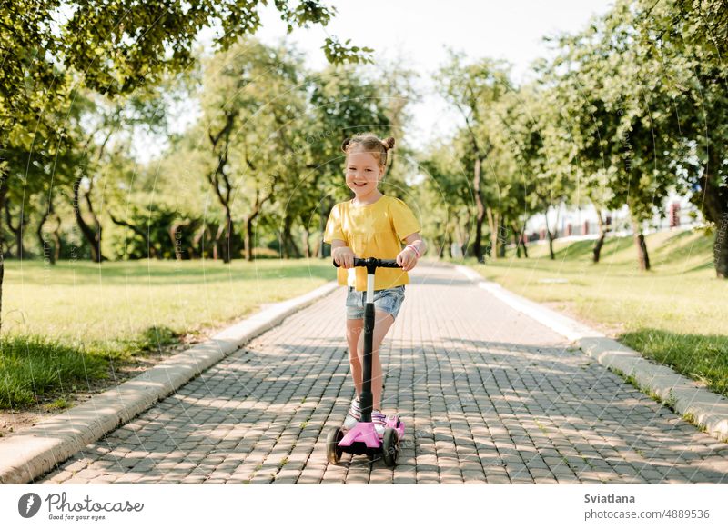 Ein aktives kleines Mädchen fährt an einem Sommertag auf einem Pfad in einem Park im Freien mit einem Motorroller. Aktiver Sport für Kinder in der Saison. Gesunder Lebensstil in der Kindheit