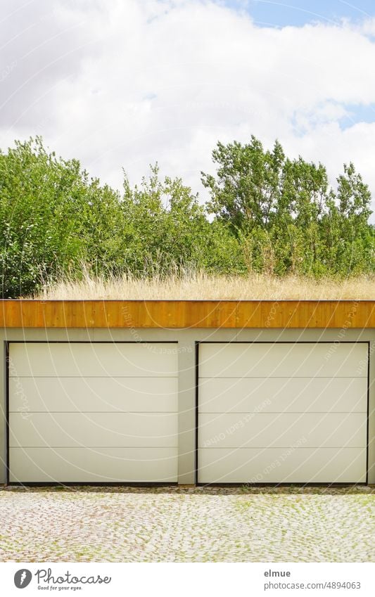 gepflasterter Platz und Doppelgarage mit vertrockneter Dachbegrünung vor grünem Buschwerk und Schönwetterwolken Garage zwei Paar paarweise Sommer Garagentor