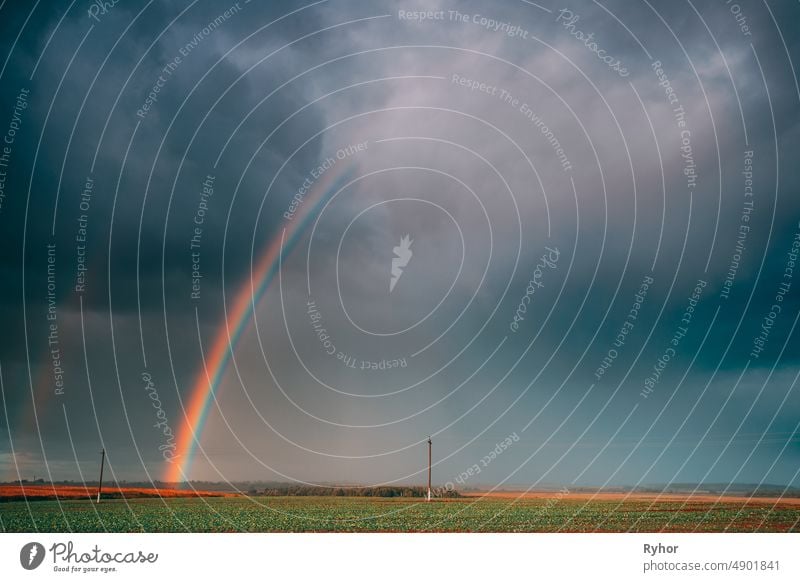 Dramatischer Himmel während des Regens mit Regenbogen am Horizont über ländlichem Landschaftsfeld. Landwirtschaft und Wettervorhersage Konzept. Ländliche Wiese im Herbst regnerischen Tag