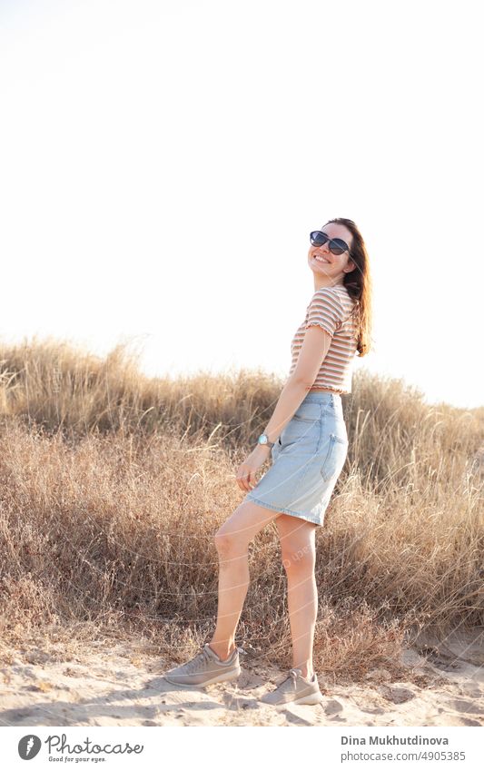 Glückliche junge Frau mit Sonnenbrille, die in der Nähe des Strandes in den Dünen mit hohem trockenem Gras spazieren geht. Foto in neutralen Beigetönen.