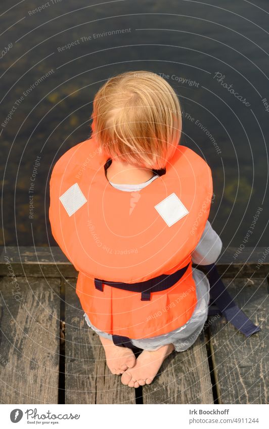 kleines Kind mit Schwimmweste hockt auf einem Steg und blickt ins Wasser im Freien sonnig tragend Meer Sommer jung Sicherheit des Lebens Kinderschwimmweste