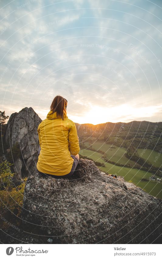 Sonnenaufgang an den Sulov-Felsen in der Ostslowakei. Schöne Brünette sitzt auf dem Felsen in gelber Jacke. Raue, unberührte Landschaft mit Felsen in orangefarbenem Licht