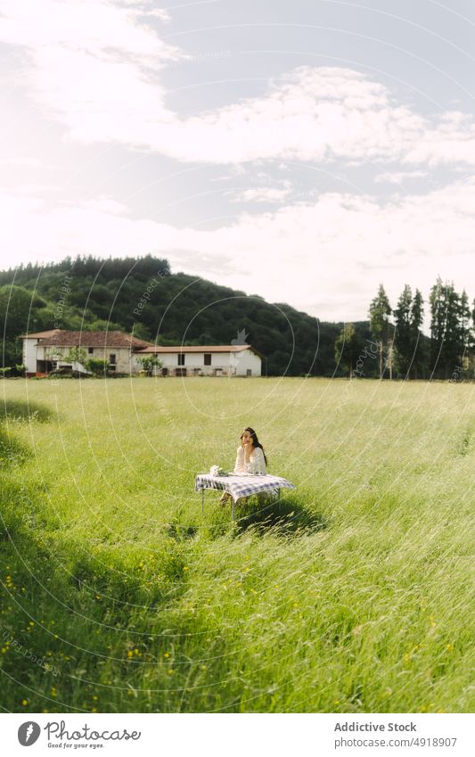 Nachdenkliche Frau sitzt am Tisch in einem Feld Picknick Landschaft Sommer Erholung Natur Gras Zeitvertreib ruhen feminin Lächeln Getränk Kleid Optimist