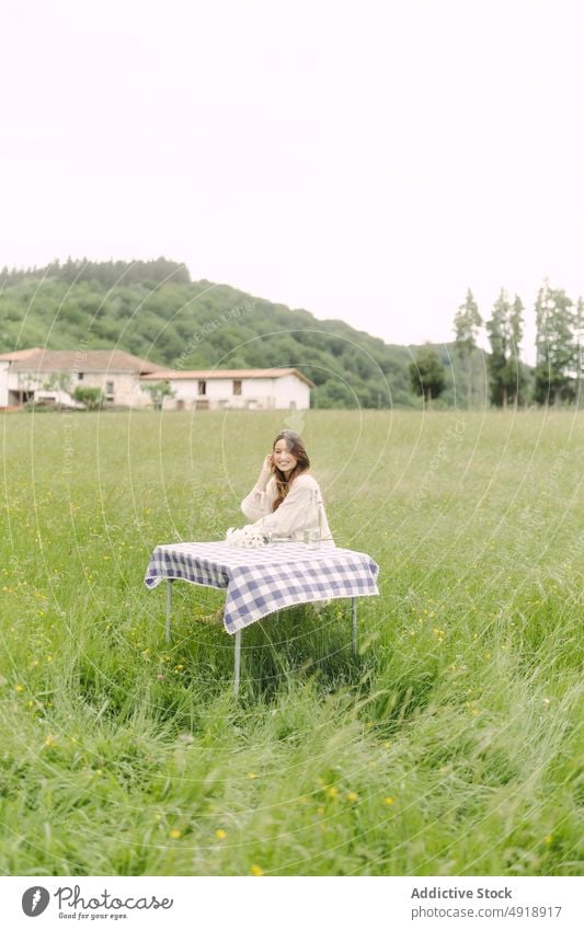 Positive Frau sitzt am Tisch in einem Feld Picknick Landschaft Sommer Erholung Natur Gras Zeitvertreib ruhen feminin Lächeln Getränk Kleid Optimist Inhalt