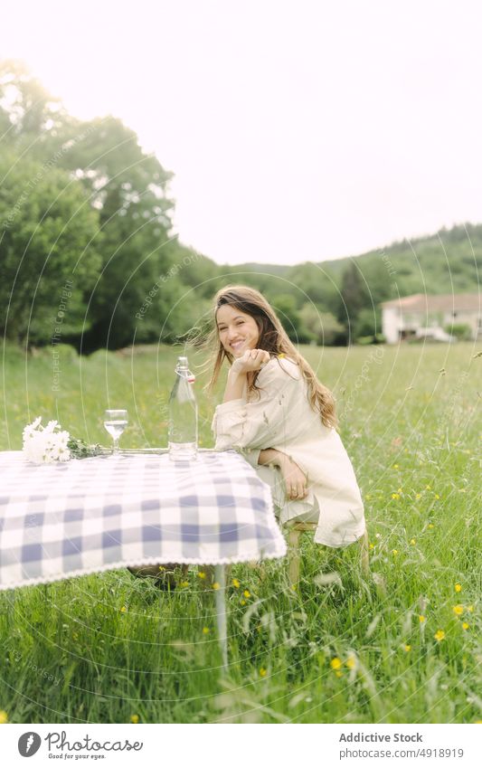 Positive Frau sitzt am Tisch in einem Feld Picknick Landschaft Sommer Erholung Natur Gras Zeitvertreib ruhen feminin Lächeln Getränk Kleid Optimist Inhalt