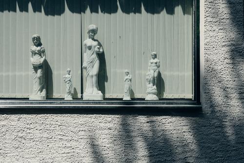 Weibliche Statuen aus Gips in einem Fenster vor Lamellen, rauh verputzte Wand und Schatten weiblich Sonne Putz Kunst griechisch kurios Standbilder Schönheit