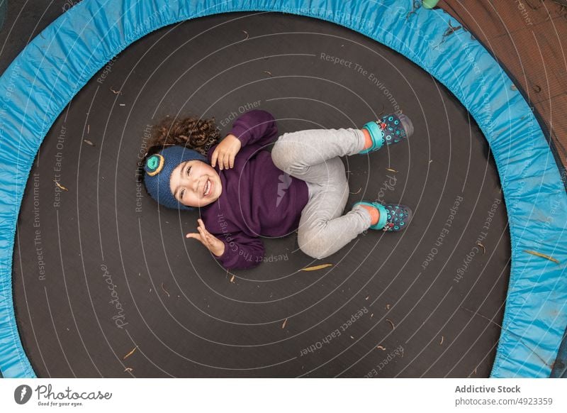 Fröhliches ethnisches Mädchen auf Trampolin liegend Kind Lügen Spaß haben Zeitvertreib unterhalten Vergnügen sorgenfrei spielerisch Kindheit aktiv Freude Glück