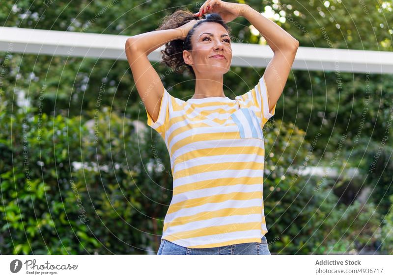 Positive Frau macht Pferdeschwanz auf der Straße Großstadt Frisur Buchse Pflanze Park Vorschein feminin Sommer heiter Lächeln grün positiv lässig Stil