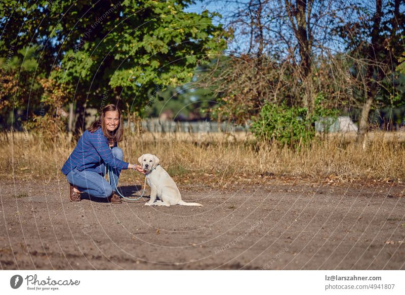 Glückliche brünette Frau mittleren Alters kniet mit ihrem kleinen Labrador-Welpen auf einem ländlichen Weg Retriever Spaziergang Leine ziehen Besitzer Sommer