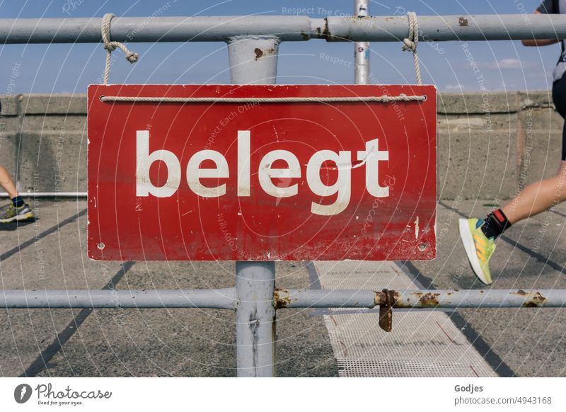 Schild mit der Aufschrift "Besetzt" an einem Geländer. Im Hintergrund die Füße von zwei Joggern Schilder & Markierungen belegt Schiene Joggen Leberfleck