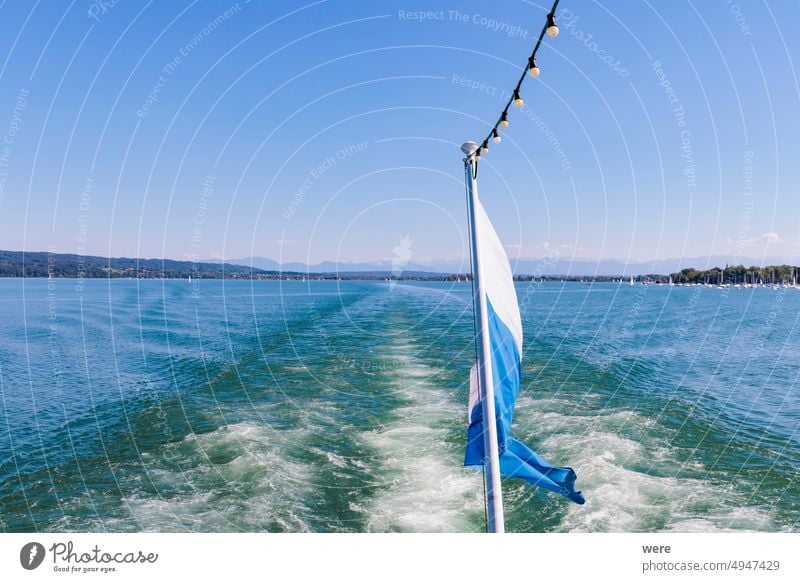 Blick von einem Schiff über die weiß-blaue bayerische Flagge auf den Ammersee in Bayern mit Segelbooten auf dem Wasser und den Ammergauer Alpen im Hintergrund