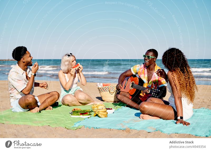 Verschiedene Freunde essen Obst und hören einem Gitarristen zu spielen Gitarre Strand zuhören Lächeln Frucht MEER Sommer Männer Frauen Wochenende Picknick Glück