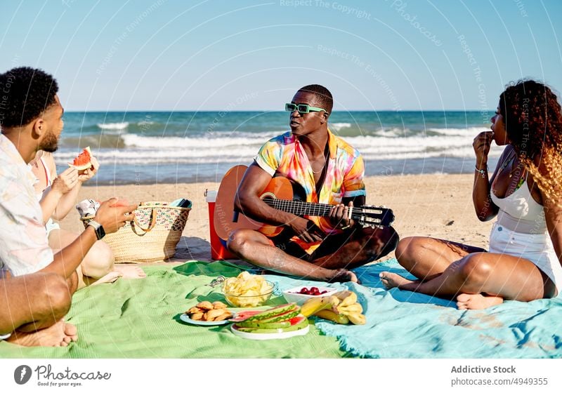 Verschiedene Freunde essen Obst und hören einem Gitarristen zu spielen Gitarre Strand zuhören Lächeln Frucht MEER Sommer Männer Frauen Wochenende Picknick Glück