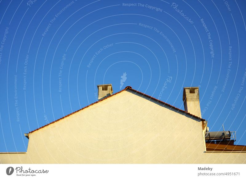 Fassade eines Wohnhaus in Beige und Naturfarben mit zwei alten Schornsteinen und Spitzgiebel vor blauem Himmel im Abendlicht in Cunda bei Ayvalik am Ägäischen Meer in Provinz Balikesir in der Türkei