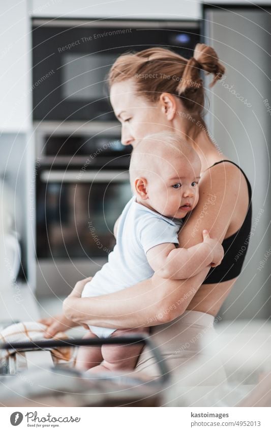 Eine Frau wischt die Spüle mit einem Tuch ab, nachdem sie den Abwasch beendet hat, während sie ein vier Monate altes Baby in ihren Händen hält. Kindheit Familie