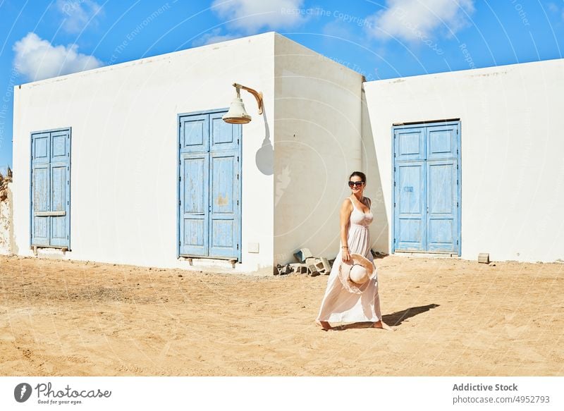 Zufriedene Frau, die vor einem weißen Haus mit blauen Fensterläden geht heiter Cottage Spaziergang Fassade Inhalt Villa Architektur Resort ländlich Sonnenkleid