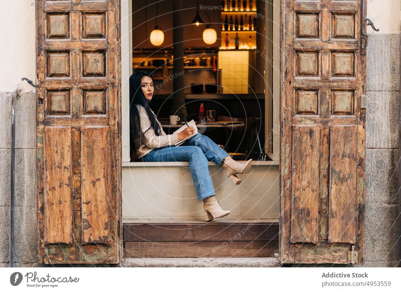 Asiatische Frau schreibt in einem Notizbuch auf der Fensterbank eines Cafés zur Kenntnis nehmen Tagebuch achtsam feminin tausendjährig charmant Schreibstift