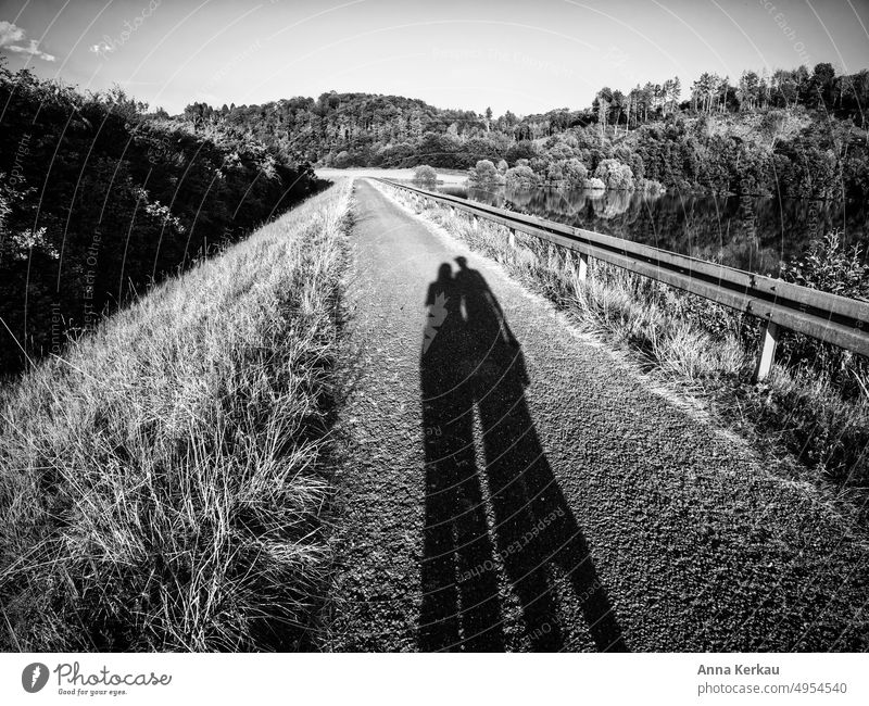 Zwei lange Schatten auf einem Asphaltweg entlang eines Seeufers Paar Silhouette zwei Personen 2 spazierengehen Licht & Schatten Zusammenhalt Partnerschaft