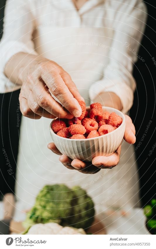 Frau hält eine Tasse mit Himbeeren und zeigt die Qualität in der Nähe up.preparing die Zutaten für eine Sauce, um das Rezept zu verbessern.black Hintergrund mit weißer Kleidung und Brokkoli auf dem Tisch