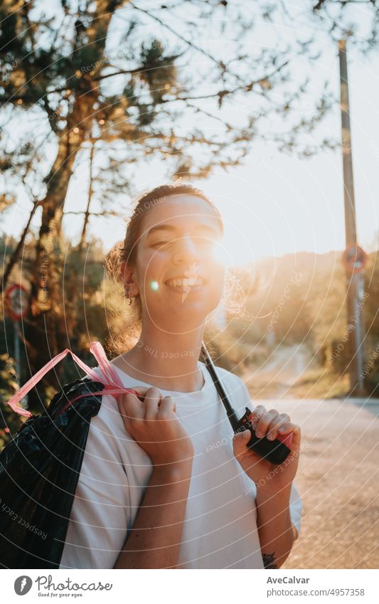 Junge Frau sammelt Müll mit einer Müllzange auf und lächelt in die Kamera.Waldhintergrund mit Sonnenuntergang, Glück.Neue Generation, die lehrt, Müll im Wald zu sammeln und zu recyceln.Bewusstsein, erneuerbare Quellen