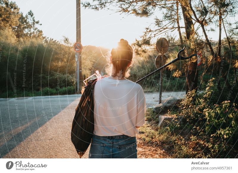 Aktivistische junge Frau, die mit einer Müllzange rückwärts zur Kamera Müll aufsammelt.Waldhintergrund mit Sonnenuntergang.Neue Generation.Freiwillige Reinigung von Wäldern und Feldern.Umwelt,Recycling,Bewusstsein,erneuerbare Quellen,Ökologie