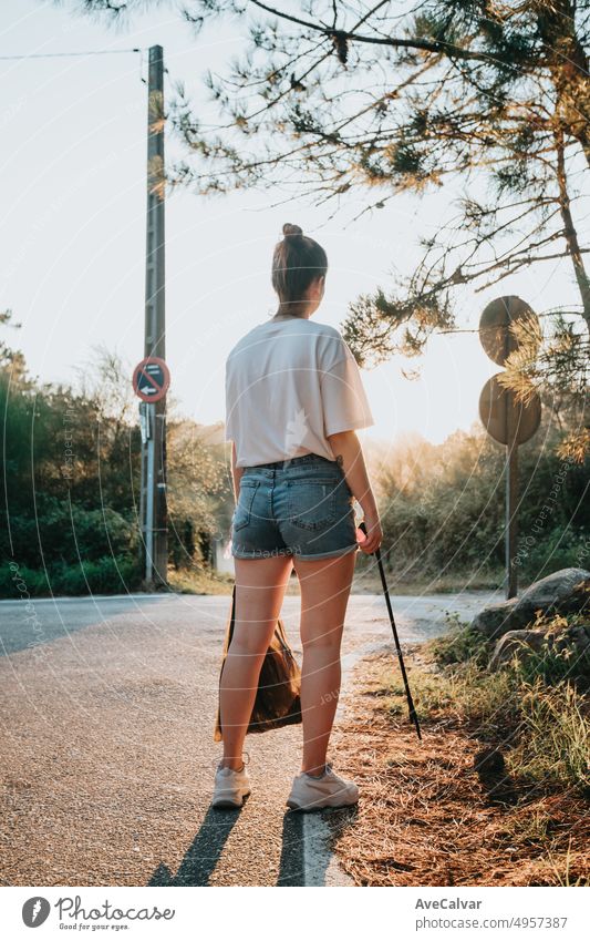 Aktivistische junge Frau, die Müll mit einer Müllzange aufhebt, rückwärts zur Kamera. Waldhintergrund mit Sonnenuntergang, friedlich, Achtsamkeit, neue Generation, Recycling, Schutz des Planeten, Umweltschutz