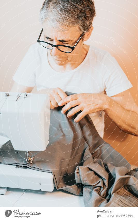 Ältere geschickte Frau näht mit faltigen Händen in einer Nähmaschine.Reparieren von Kleidung.Hose,Hemd,Rock,Kleid.Grauer Stoff und Nadel.Oma lehrt, wie man auf sozialen Medien näht