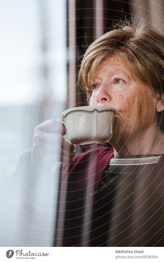 Nachdenkliche ältere Frau trinkt heißes Getränk am Fenster gemütlich trinken Tasse heimwärts besinnlich ruhen in den Ruhestand gehen lässig Raum Lifestyle