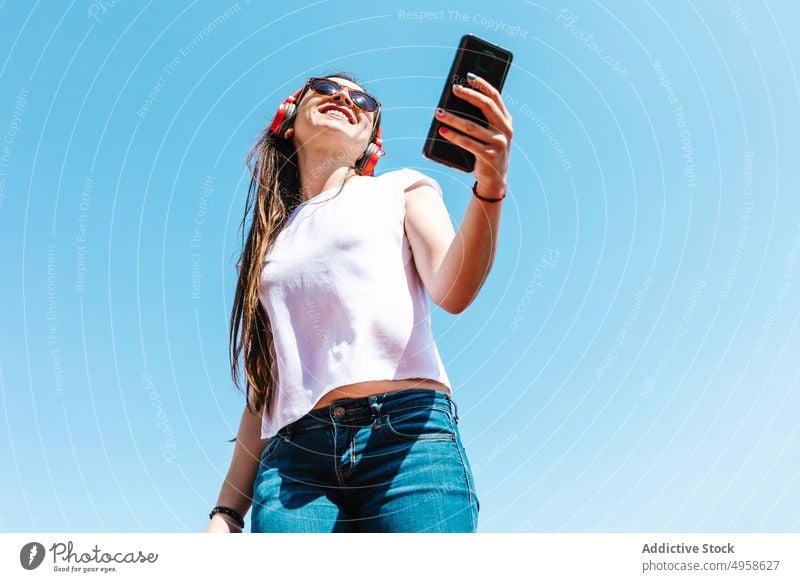 Ethnische Frau mit Sonnenbrille und Smartphone im Sonnenlicht Stil heiter Individualität Glück Kopfhörer Lifestyle benutzend Apparatur Gerät Zeitgenosse Design