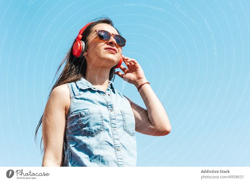 Ethnische Frau mit Sonnenbrille und drahtlosen Kopfhörern im Sonnenlicht Stil heiter Individualität Gesang Glück Klang meloman Lifestyle Headset benutzend