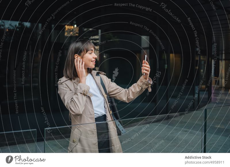 Fröhliche Frau mit Videoanruf in der Nähe eines modernen Gebäudes Kopfhörer Smartphone Video-Chat Kommunizieren Gespräch Straße urban trendy Terrasse Outfit