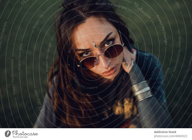 Ruhige flippige Millennial-Frau auf Rasen im Park Tattoo Verlockung Vorschein brünett Schmuck Sonnenbrille cool Piercing Windstille Accessoire provokant Gras