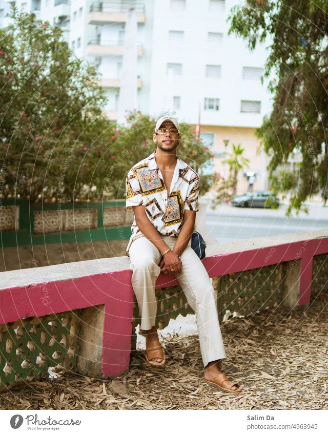 Stilvoll lächelnder Mann im Park sitzend stylisch männlich Hut Verschlussdeckel Stilrichtung trendy Mode modisch Blick Blick in die Kamera Pose posierend Model