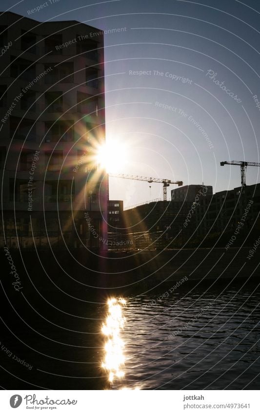 Sonnenstrahlen hinter einem modernen, mehrstöckigen Haus, daneben Baukräne, im Vordergrund Wasser Berlin Baustelle Gebäude Architektur Wohnhaus Mehrfamilienhaus