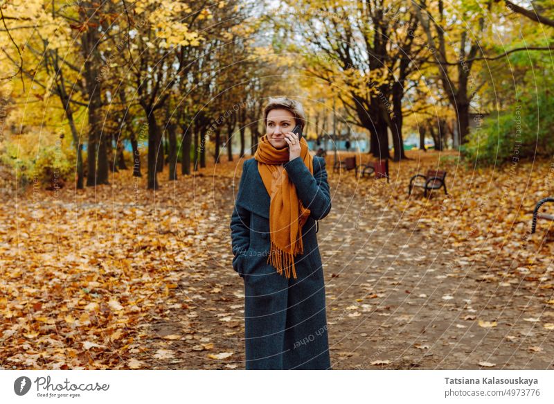 Eine Blondine in Mantel und Schal telefoniert, während sie durch einen herbstlichen Park geht. Frau fallen Herbst Anruf Telefon Mobile Handy Mitteilung