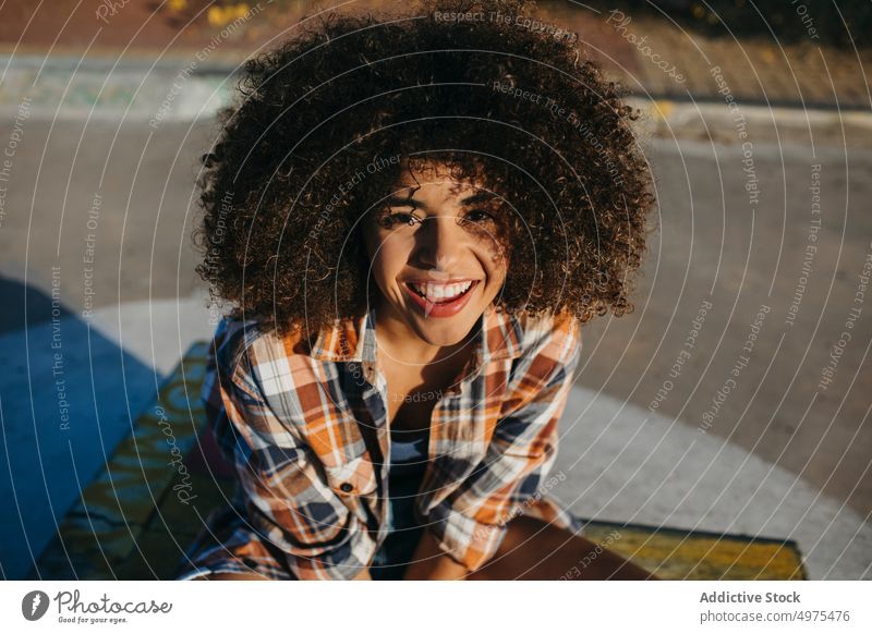 Charmante schwarze Frau mit lockigem Haar lächelnd auf der Straße Hipster Sonnenuntergang Afro-Look Frisur Inhalt charmant Lächeln kariertes Hemd ethnisch