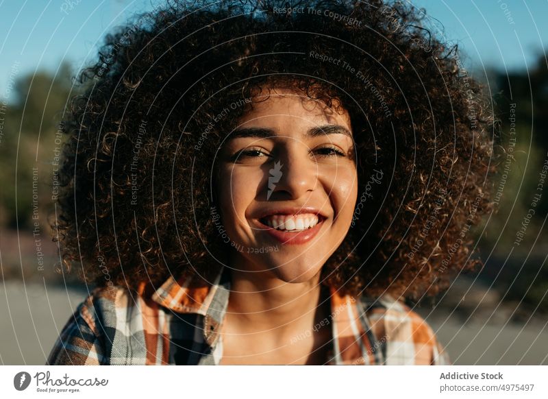 Charmante schwarze Frau mit lockigem Haar lächelnd auf der Straße Hipster Sonnenuntergang Afro-Look Frisur Inhalt charmant Lächeln kariertes Hemd ethnisch