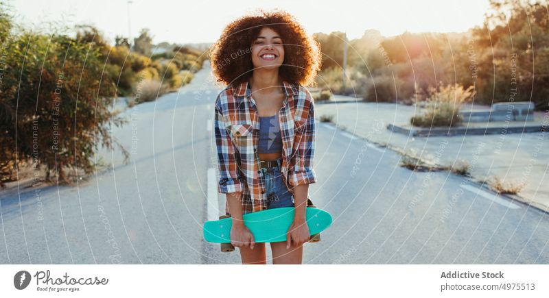 Fröhliche ethnische Frau mit Penny Board Lochrasterplatte Straße Hipster Sommer Skater Afro-Look Sonnenuntergang heiter tausendjährig Afroamerikaner schwarz