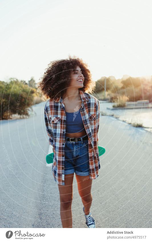 Fröhliche ethnische Frau mit Penny Board Lochrasterplatte Straße Hipster Sommer Skater Afro-Look Sonnenuntergang heiter tausendjährig Afroamerikaner schwarz