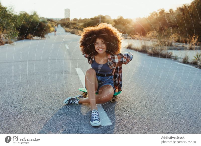 Entzückende afroamerikanische Frau, die auf einem Penny Board auf der Asphaltstraße sitzt Lochrasterplatte Sommer Skater Straße cool tausendjährig Afro-Look