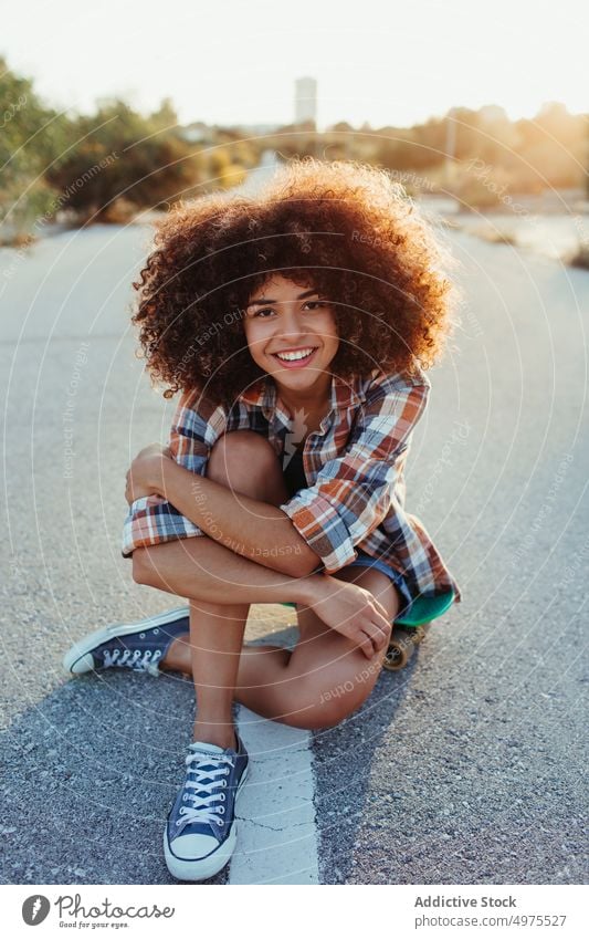 Entzückende afroamerikanische Frau, die auf einem Penny Board auf der Asphaltstraße sitzt Lochrasterplatte Sommer Skater Straße cool tausendjährig Afro-Look