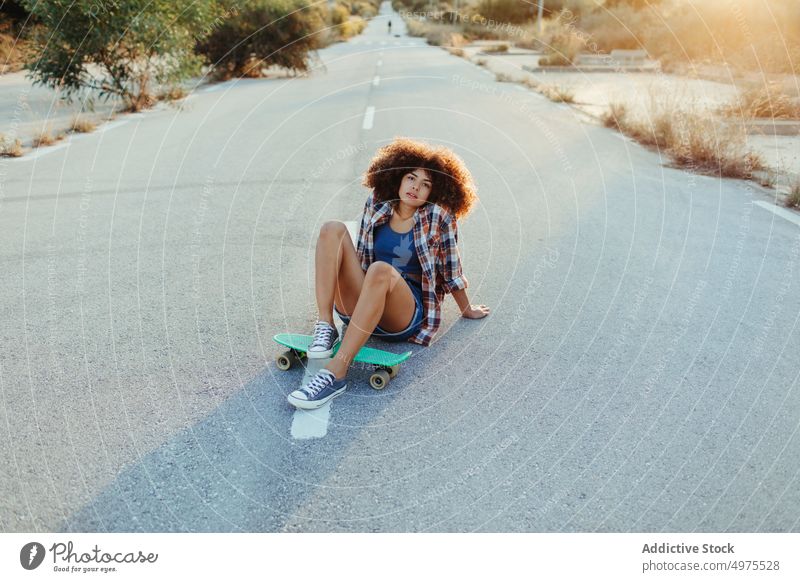 Afroamerikanische Frau sitzt auf einem Penny Board auf der asphaltierten Straße Lochrasterplatte Sommer Skater cool tausendjährig Afro-Look Asphalt trendy ruhig