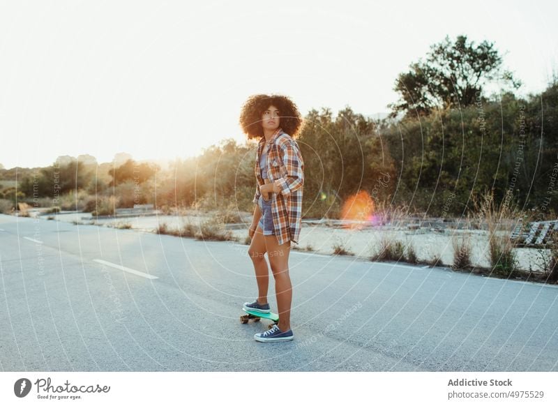 Ethnische Frau reitet Penny Board Lochrasterplatte Mitfahrgelegenheit Sonnenuntergang Straße Sommer Hipster Skater Afro-Look urban ethnisch trendy ruhig