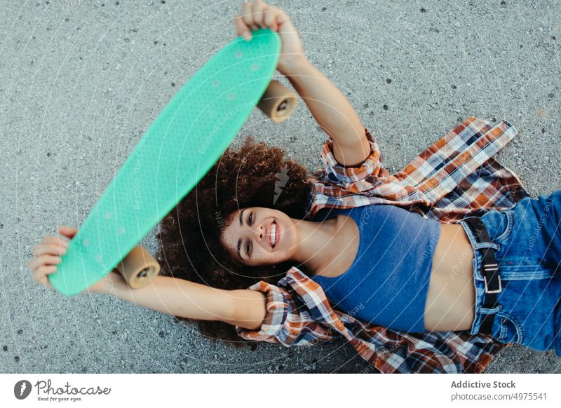 Eine afroamerikanische Frau liegt mit einem Brett auf der asphaltierten Straße Lochrasterplatte Sommer Skater tausendjährig Afro-Look Asphalt trendy ruhig