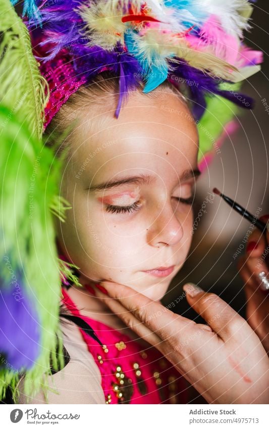 Crop Mutter Anwendung Festival Make-up auf Tochter Gesicht Karneval vorbereiten Verkehrsflugzeug bewerben heimwärts farbenfroh Feder Kopfbedeckung Mädchen Frau