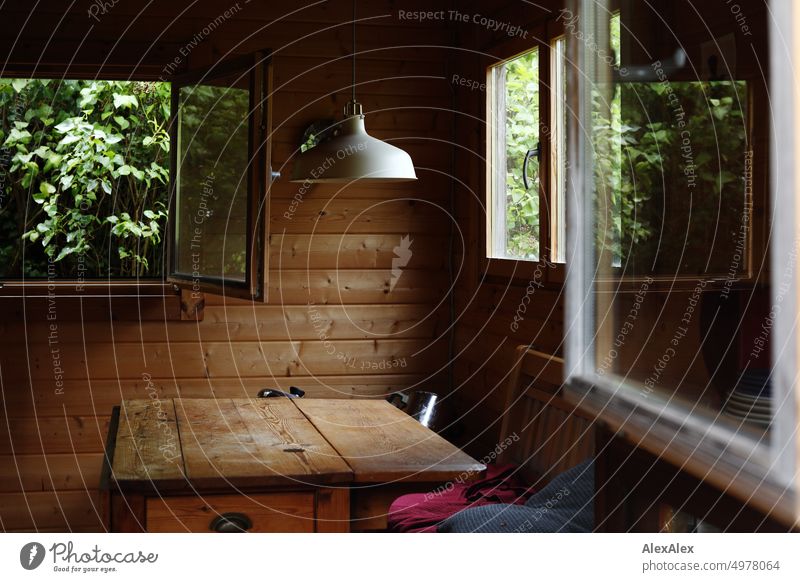 Einblick durch geöffnetes Fenster in eine Gartenhütte aus Holz mit Holztisch, Bank und anderen Fenstern. Gartenlaube Gartenhäuschen Hütte Bungalow Tisch Lampe