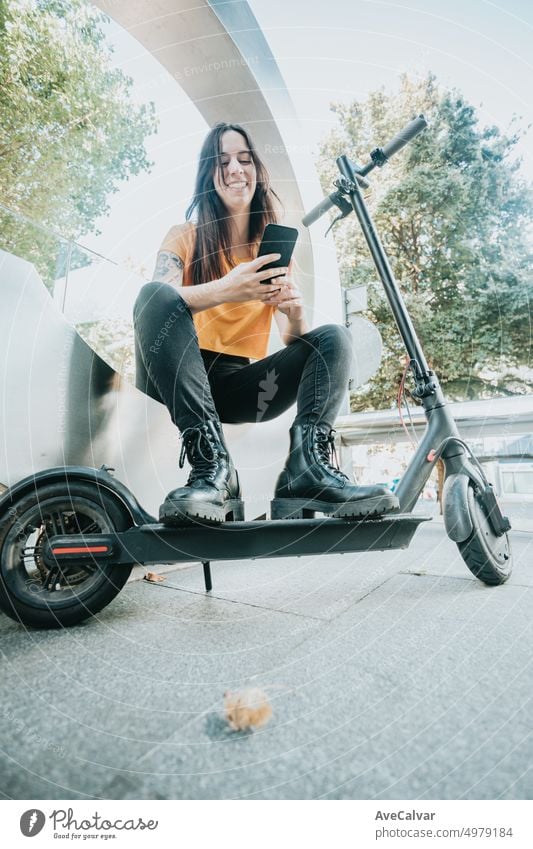 Porträt einer jungen Frau, die in einer städtischen Landschaft sitzt, ein Handy hält und sich mit einem Lächeln auf einen Elektroroller stützt. Ausruhen und beobachten soziale Medien mit lässiger Kleidung. Stadt Lebensstil