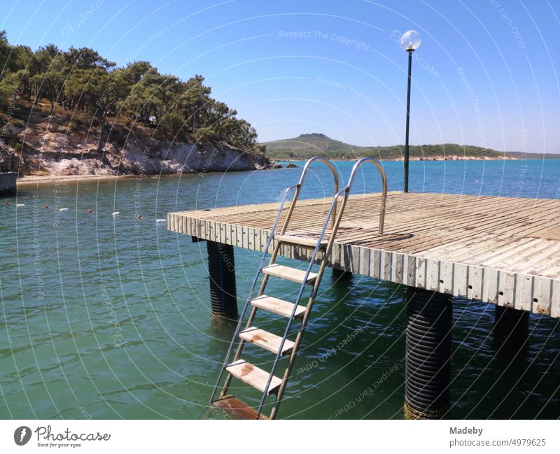 Bootssteg mit Schiffsdielen und Badeleiter im Sommer bei blauem Himmel und Sonnenschein am Strand von Ayvalik am Ägäischen Meer in der Provinz Izmir in der Türkei