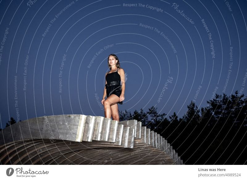 Der Himmel ist dunkelblau. Ein wunderschönes brünettes Mädchen ist in Schwarz gekleidet. Sie schaut direkt in eine Kamera. Sie ist braungebrannt und modisch. Eine hübsche Frau und ihr Model-Test vor Ihnen.