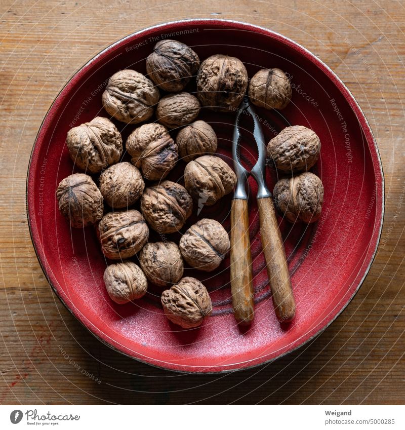 Runde, rote Keramikschale auf einem Holztisch zentral von oben fotografiert, in der Walnüsse und ein Nussknacker mit unregelmäßigem Holzgriff liegen